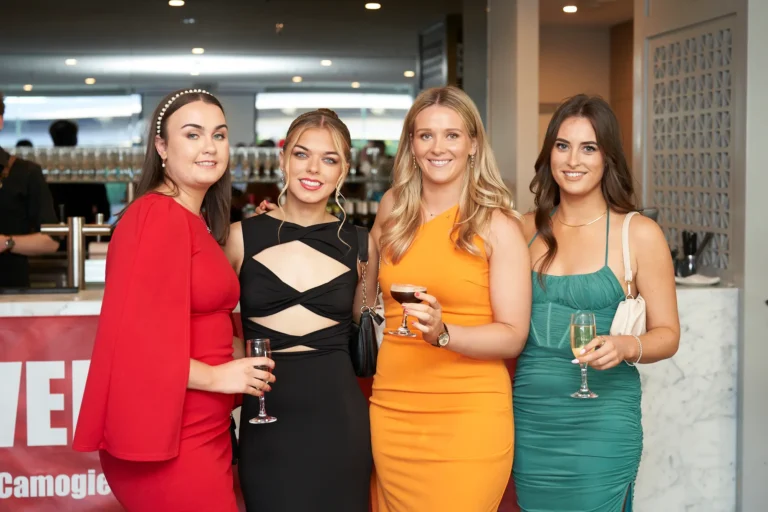 Four women in formal attire smiling and holding drinks while posing together at the bar during the Garryowen Awards Night at River’s Edge Events