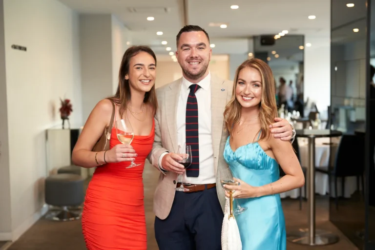 Three smiling individuals guests at River’s Edge Events during the Garryowen Awards Night, two women and one man, stand together holding drinks in a bright, indoor event space. The man in the center wears a suit jacket and striped tie, flanked by a woman in a red dress on the left and a woman in a blue dress on the right. Both women are holding wine glasses