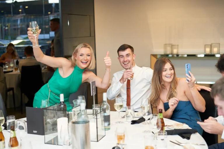 garryowen awards night rivers edge events guests table candid Three smiling people at River’s Edge Events during the Garryowen Awards Night: a blonde woman in a green dress raises a wine glass, a man in a white shirt and tie gives a thumbs-up while holding a drink, and a woman in a blue dress laughs while holding up her phone as if taking a selfie or video. They are seated at a table with bottles and glasses
