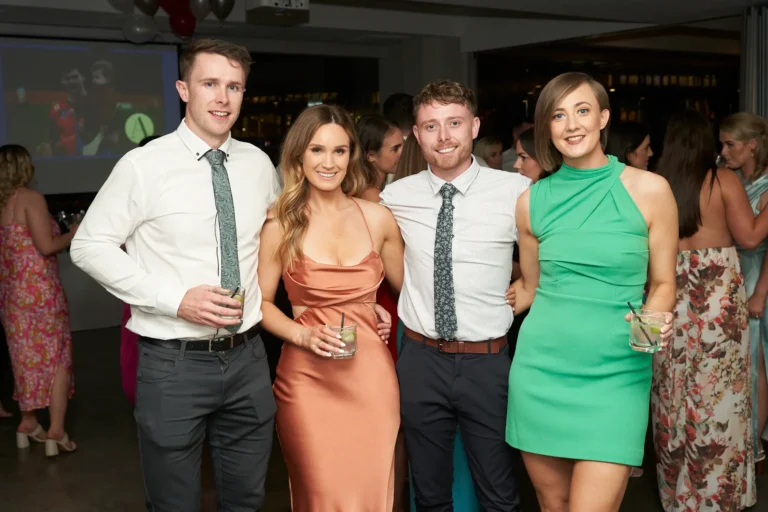 Four people, two men in dress shirts and ties and two women in peach and green dresses, smiling and holding drinks at the Garryowen Awards Night at River’s Edge Events, with other guests blurred in the dimly lit background