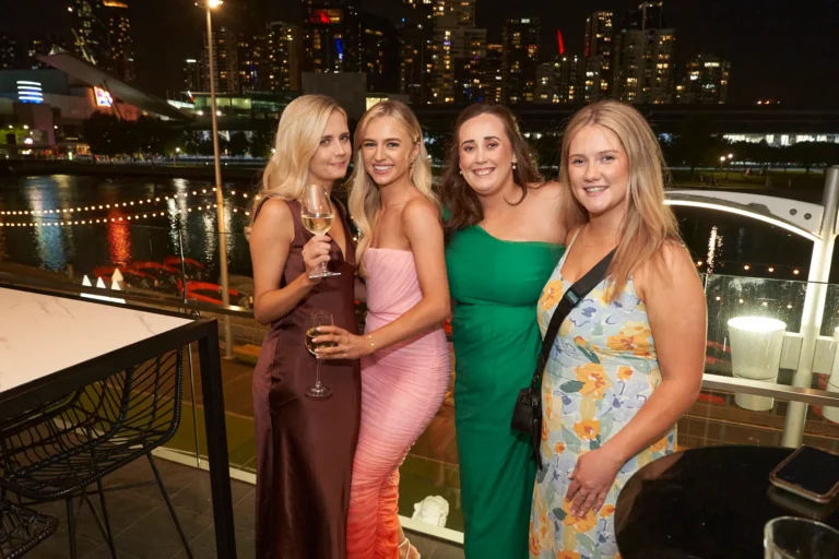 Four women in formal dresses posing and smiling on an outdoor balcony at night during the Garryowen Awards Night at River’s Edge Events, with Melbourne city lights and the Yarra River in the background