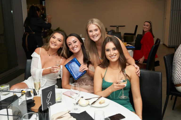 Four women smiling and posing at a table during a formal dinner at the Garryowen Awards Night at River’s Edge Events, holding drinks and one holding an award