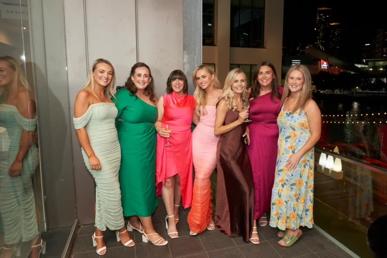 garryowen awards night rivers edge group seven women balcony An image of seven young women posing together at a formal event at the Garryowen Awards Night at River’s Edge Events. They are dressed in various formal gowns in shades of green, pink, orange, brown, and floral patterns. They are standing outdoors on a balcony, with city lights and waterfront visible in the background at night. The women are smiling and enjoying the event