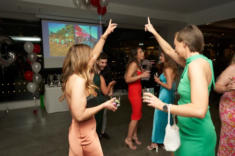 A group of formally dressed guests celebrating and dancing with balloons and trophies in the background, and a projected image on a screen at the Garryowen Awards Night at River’s Edge Events