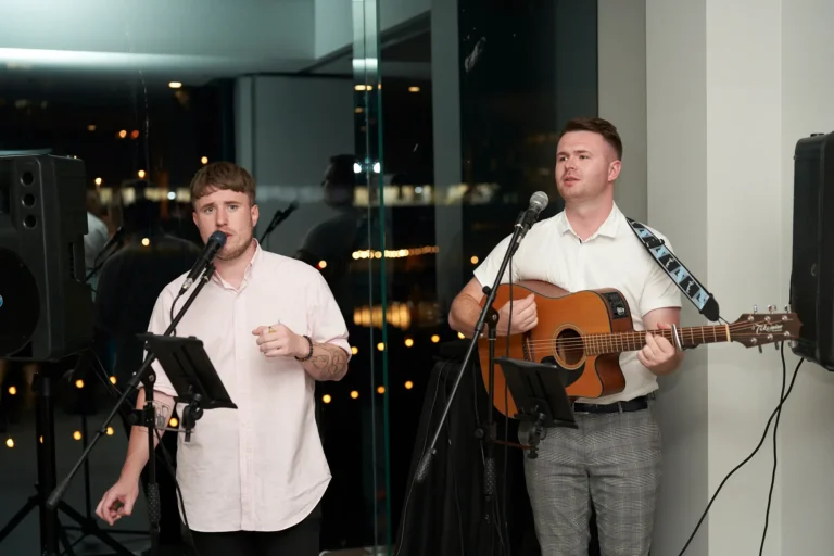 Two men performing live music indoors at night, one singing into a microphone and the other singing while playing an acoustic guitar, at the Garryowen Awards Night at River’s Edge Events, with speakers and music stands visible