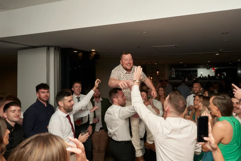 A man in a floral shirt being lifted overhead by several others on a crowded dance floor, surrounded by cheering and smiling guests during the Garryowen Awards Night at River’s Edge Events, capturing an energetic and celebratory moment