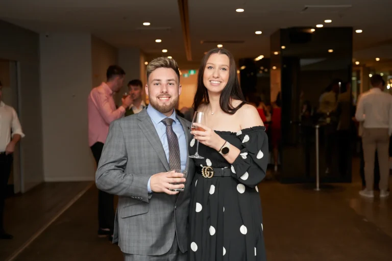 A man in a grey suit and a woman in a black off-the-shoulder polka dot dress standing side-by-side and holding drinks, smiling at the Garryowen Awards Night at River’s Edge Events, with other attendees visible in the dimly lit background