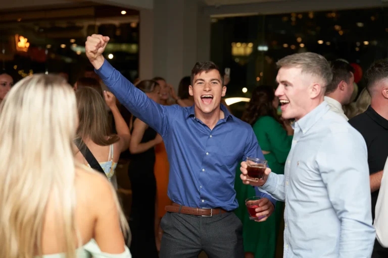 An excited man in a blue shirt raises his fist in celebration while another man holding a drink laughs at the Garryowen Awards Night at River’s Edge Events, with guests dancing and city lights visible through the windows in the background