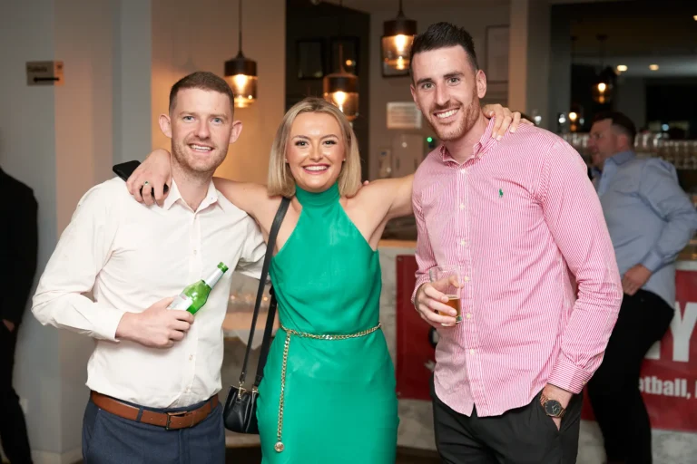 A group of three smiling friends, two men and one woman, are posing with their arms around each other at the Garryowen Awards Night at River’s Edge Events. The man on the left wears a white shirt and holds a green bottle; the woman in the center wears a vibrant green halter-neck dress with a gold chain belt; and the man on the right wears a pink striped shirt and holds a drink in a glass. They appear happy and relaxed in an indoor setting with a bar and blurred background details