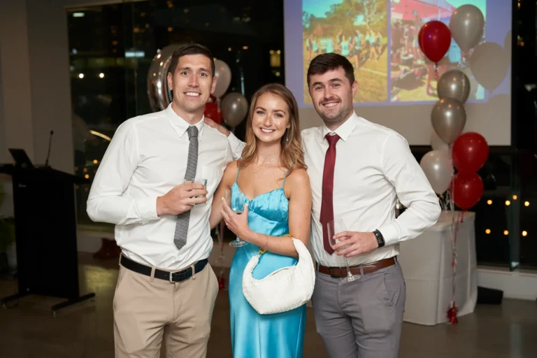 Three formally dressed individuals—two men in white shirts and ties and a woman in a light blue satin dress holding a white handbag—posing at the Garryowen Awards Night at River’s Edge Events, with balloons and a projected image in the background