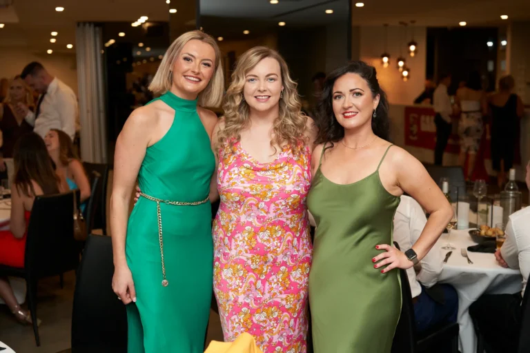 Three women in formal dresses—a green halter-neck dress with gold chain belt, a pink and orange floral print dress, and a green satin slip dress—posing and smiling at the Garryowen Awards Night at River’s Edge Events, with tables and other attendees in the dimly lit background