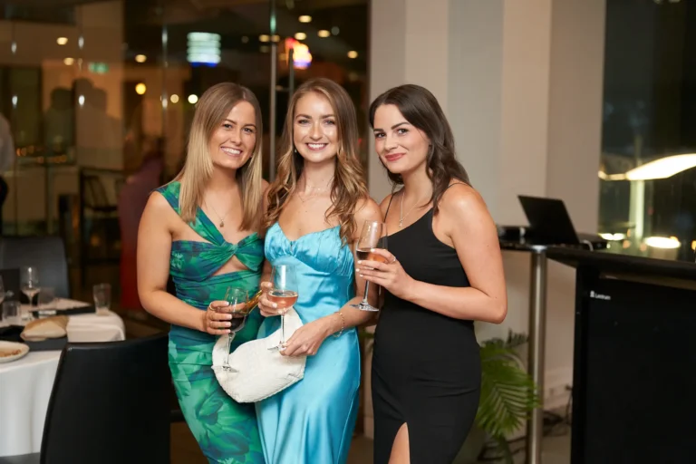 Three women in formal attire—a green patterned dress, a light blue satin dress, and a black dress with a slit—holding drinks and smiling at the Garryowen Awards Night at River’s Edge Events, standing in front of a glass wall with tables and chairs in the background
