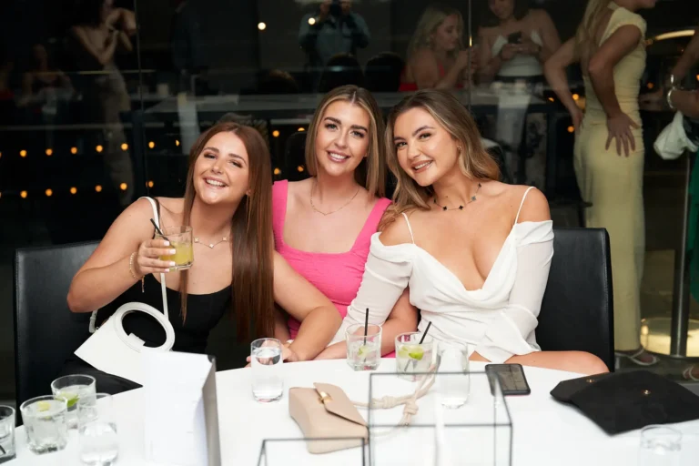 Three smiling young women sit at a table in a dimly lit. The woman on the left, with long brown hair, wears a black top and holds a drink. The woman in the middle, with blonde hair, wears a bright pink top. The woman on the right, also with blonde hair, wears a white off-the-shoulder top. Drinks are on the table in front of them, and other attendees are visible blurred in the background