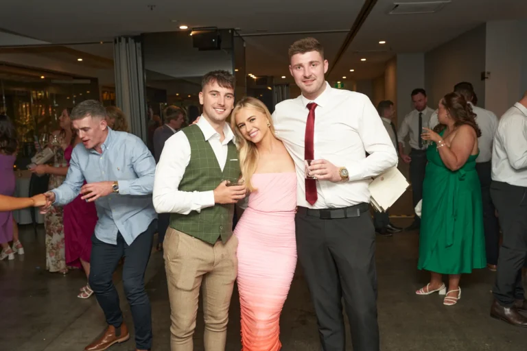 Three young adults are smiling at the camera at the Garryowen Awards Night at River’s Edge Events. A man on the left wears a green vest over a white shirt and tan trousers, while a woman in the center wears a light pink, fitted dress. A tall man on the right wears a white shirt, a red tie, and dark trousers. Other guests are visible in the blurred background of a dimly lit venue