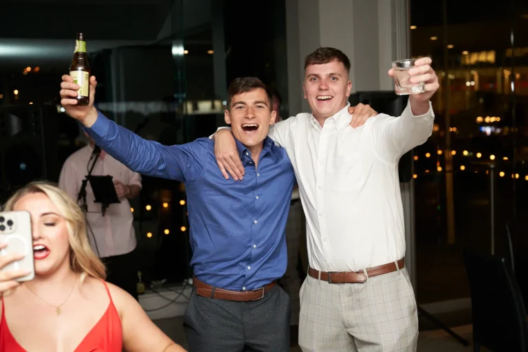 Two smiling men raising their arms and holding drinks while celebrating at the Garryowen Awards Night at River’s Edge Events, with a woman in the foreground taking a photo with her phone
