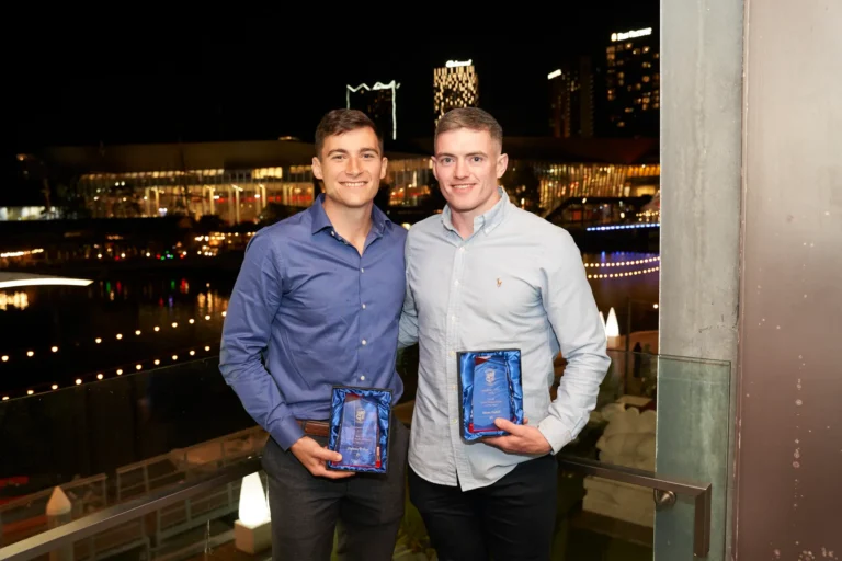 Two men smiling and holding awards on an outdoor balcony at night during the Garryowen Awards Night at River’s Edge Events, with Melbourne city skyline and the Yarra River visible in the background