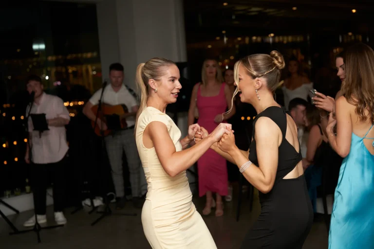Two women smiling and holding hands while dancing at a lively evening event at the Garryowen Awards Night at River’s Edge Events, with other guests and musicians in the dimly lit background and city lights visible through the glass wall