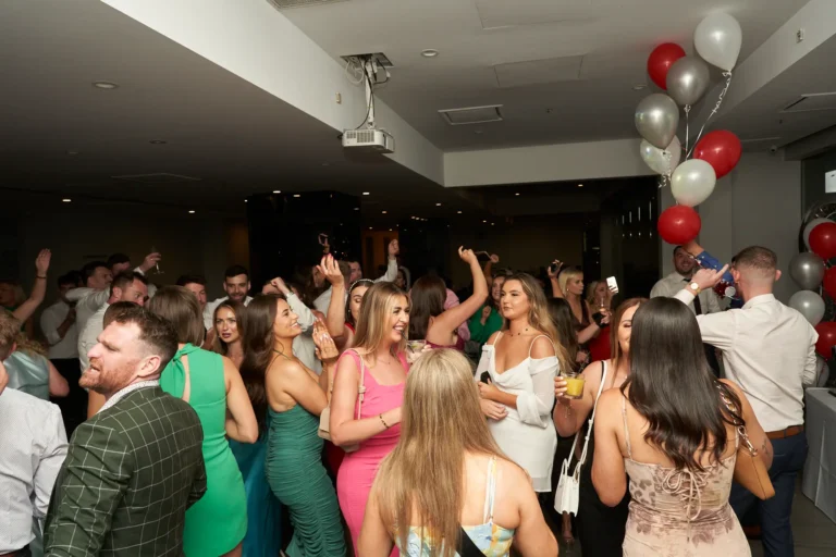 A lively wide shot of a large group of well-dressed guests dancing on the dance floor, including a man in a green and black checkered suit in the foreground, at the Garryowen Awards Night at River’s Edge Events, with balloons and drinks visible in the background