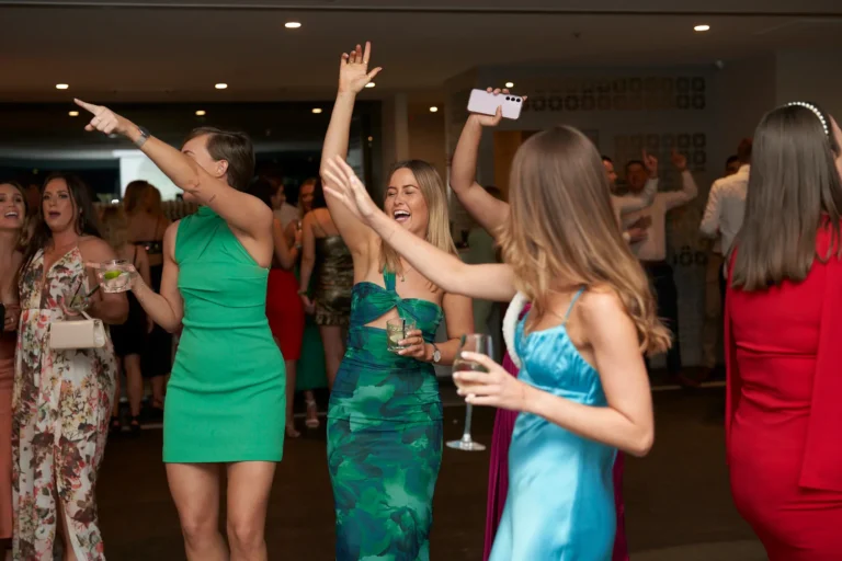 A group of women in festive dresses celebrating indoors, some dancing and holding drinks, with a woman in a vibrant green dress laughing with her arms raised at the Garryowen Awards Night at River’s Edge Events