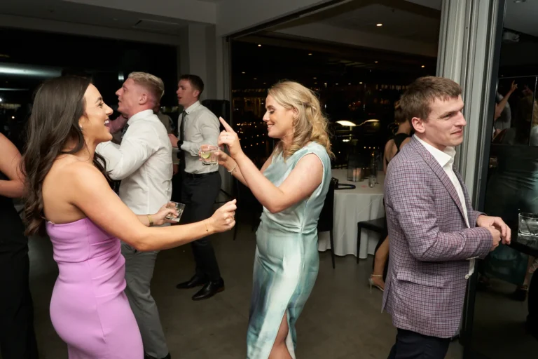 Two women dancing and smiling in the foreground while a group of well-dressed guests socialize and dance in the background at the Garryowen Awards Night at River’s Edge Events, with large glass doors showing the outdoor area at night