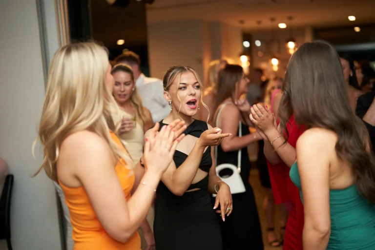 A group of young women dancing at a lively event, with a woman in a black cutout dress animatedly speaking with her hands toward a woman in an orange dress, at the Garryowen Awards Night at River’s Edge Events