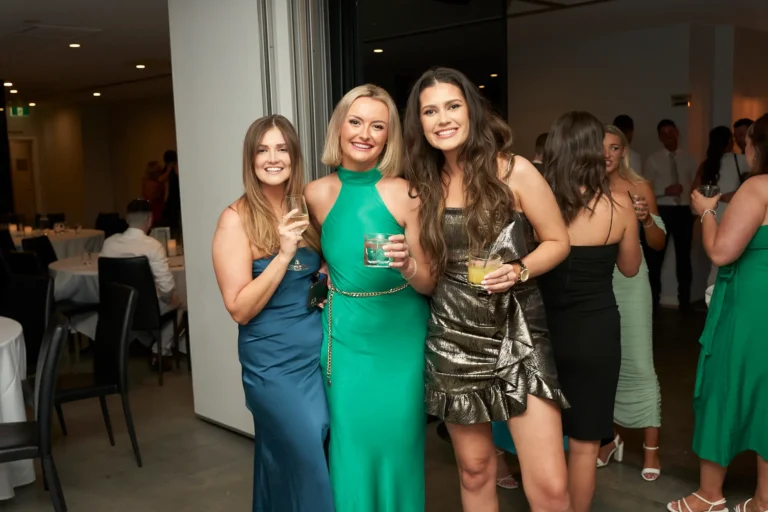 Three women in formal dresses—a blue maxi dress, a green halter-neck maxi dress, and a gold mini-dress—smiling with drinks at the Garryowen Awards Night at River’s Edge Events, with other guests and tables in the background