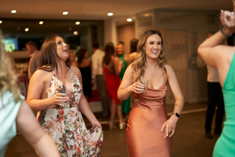 Two women holding drinks—one in a floral dress and the other in a copper-colored dress—laughing and dancing joyfully at the Garryowen Awards Night at River’s Edge Events, with other guests dancing in the dimly lit background