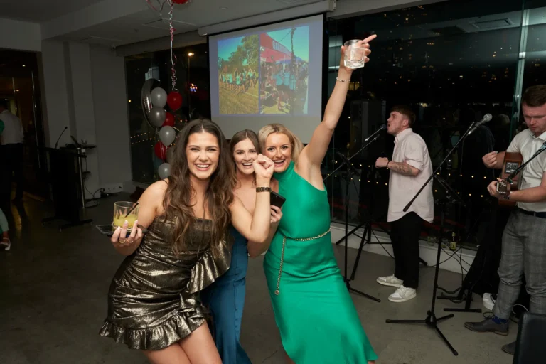 Three smiling women pose with drinks, with a band performing in the background and a projection screen displaying an outdoor scene. One woman in a shimmering gold dress holds a drink, another in blue laughs, and a third in a green dress raises her drink in celebration. Balloons are visible in the background
