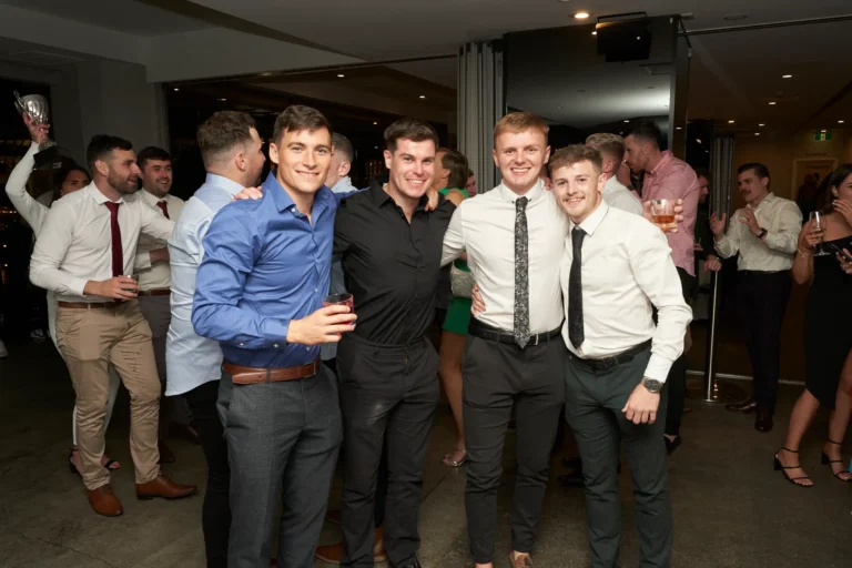 An indoor scene showing a group of young men at the Garryowen Awards Night. Four men in the foreground are smiling and posing for the camera, with the two central figures having their arms around each other. They are dressed in smart casual attire, including collared shirts, ties, and trousers. Other attendees are visible in the background, also engaged in conversation and holding drinks, suggesting a lively atmosphere