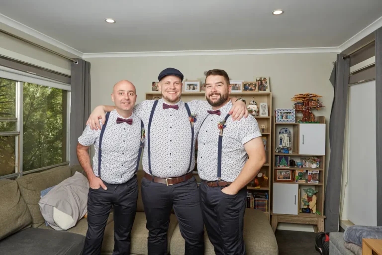 Groom with arm around groomsman’s shoulders, both smiling indoors during groom’s preparation, groomsman with hand in pocket