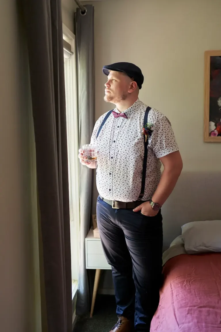 Groom standing near window looking outside, holding glass of whisky with one hand and other hand in pocket indoors during groom’s preparation
