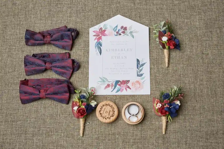 Styling shot of boutonnière, wedding rings, bow tie, and invitation card during groom’s preparation