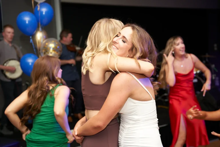 Candid moment at St. Kevin’s 65th Anniversary Ball featuring two women warmly embracing, one in a brown outfit and the other in a white spaghetti-strap top smiling with eyes closed. In the background, a woman in a red dress dances while musicians perform with instruments under festive blue and gold balloons, capturing the joyous spirit of the celebration.