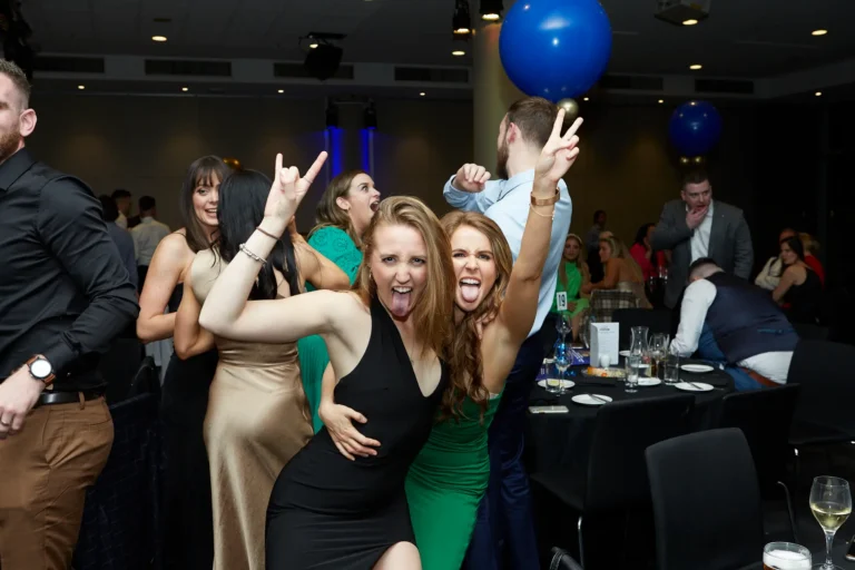 Candid moment at St. Kevin’s 65th Anniversary Ball showing two women in elegant dresses making playful "rock on" gestures and sticking out their tongues, capturing the fun and lively spirit of the evening. In the background, other guests socialize, dance, and raise their hands in excitement, while blue balloons and tables set for dinner add to the celebratory ballroom atmosphere.