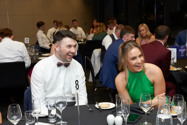 Candid moment at St. Kevin’s 65th Anniversary Ball showing a man in a white shirt with a bow tie and a woman in a green dress laughing together at a table with formal place settings. Other guests in the softly lit background add to the lively, celebratory atmosphere.