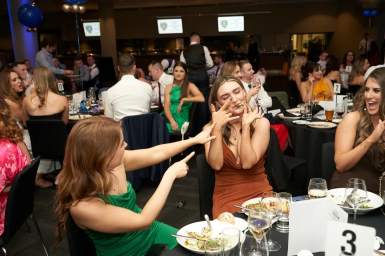 Candid moment at St. Kevin’s 65th Anniversary Ball with two women sharing laughter at a guest table. One in a green dress points and laughs, while another in a rust-colored dress playfully makes a face with her hands. The table is set with plates, glasses, and dessert, while other guests in the background socialize in the elegant, balloon-decorated venue.