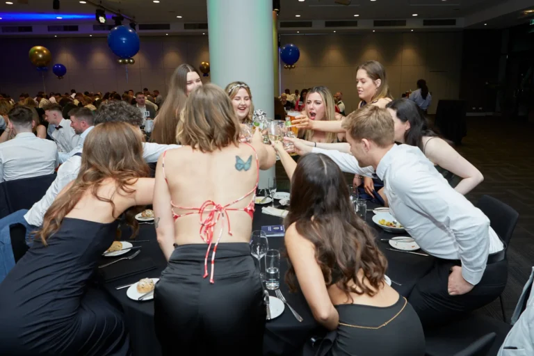 Candid moment at St. Kevin’s 65th Anniversary Ball featuring elegantly dressed guests raising their glasses in a celebratory toast around a round table. Young women in formal dresses and men in white shirts share laughter and camaraderie, with blue and gold balloons in the background adding to the festive atmosphere.