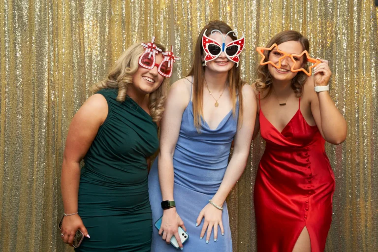 Candid moment at St. Kevin’s 65th Anniversary Ball with three young women posing playfully in front of a glittering gold sequin backdrop. One wears a dark green ruched dress holding oversized red martini-glass props, another in a light blue slip dress sports red glitter butterfly sunglasses, and the third in a vibrant red satin dress holds orange star-shaped glasses. Their joyful expressions and playful props capture the fun, celebratory spirit of the anniversary event.