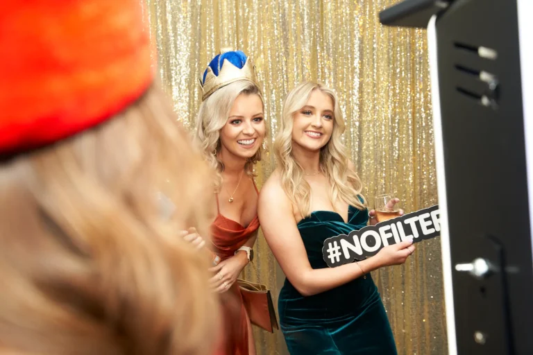 Candid moment at St. Kevin’s 65th Anniversary Ball featuring two glamorous guests posing in front of a shimmering gold backdrop. One wears a copper-toned dress with a gold and blue crown, while the other in an emerald green gown holds a "#NOFILTER" sign and a drink. Both smile broadly, capturing the joyful, festive spirit of the anniversary celebration.