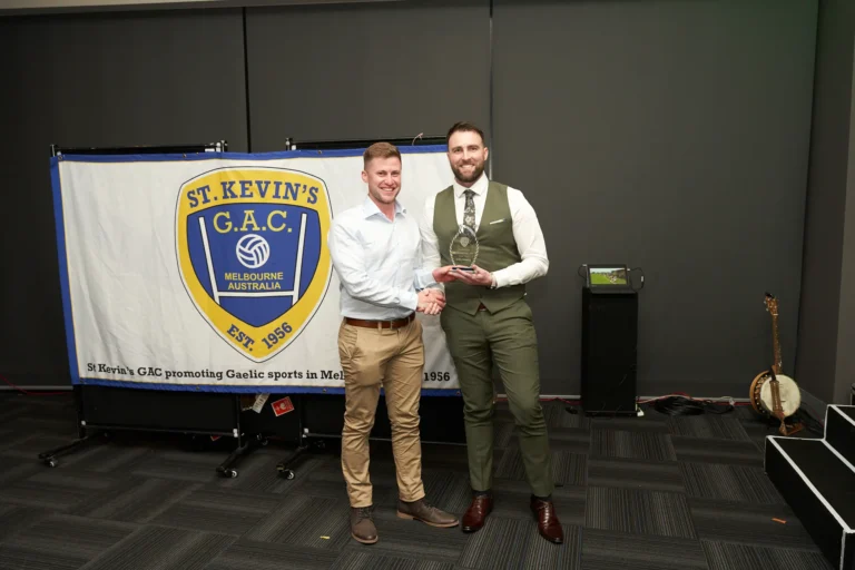 Two men share a handshake during an award presentation at St. Kevin’s 65th Anniversary Ball at Marvel Stadium, Melbourne. The recipient, wearing a green waistcoat and trousers, holds a clear trophy while the presenter, dressed in a collared shirt and chinos, congratulates him. Behind them, a banner displays the St. Kevin’s G.A.C. crest and highlights their role in promoting Gaelic sports. Guests’ tables and festive decorations create a warm, celebratory atmosphere, capturing the spirit of recognition and achievement at the milestone anniversary event.