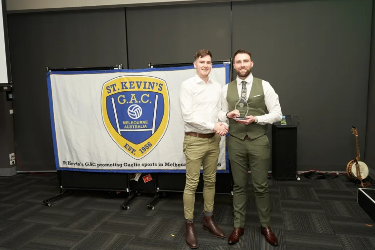 Award presentation at St. Kevin’s 65th Anniversary Ball, Marvel Stadium, Melbourne. An award recipient in a white shirt and khaki trousers shakes hands with the presenter in a dark vest and tie, while a large banner behind them displays the St. Kevin’s G.A.C. crest and the club’s mission. Guests and the formal indoor setting highlight the celebratory and prestigious atmosphere of the milestone event.