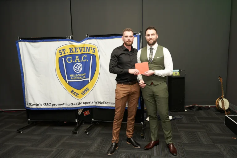 This image from St. Kevin’s G.A.C. Melbourne’s 65th Anniversary Ball at Marvel Stadium captures a formal award presentation. In the foreground, a man in a dark shirt and brown trousers receives a red envelope or award certificate from another man dressed in a green waistcoat and tie. Behind them, a large banner displays the club’s logo and “EST. 1956,” reinforcing the heritage and celebratory context of the milestone event.