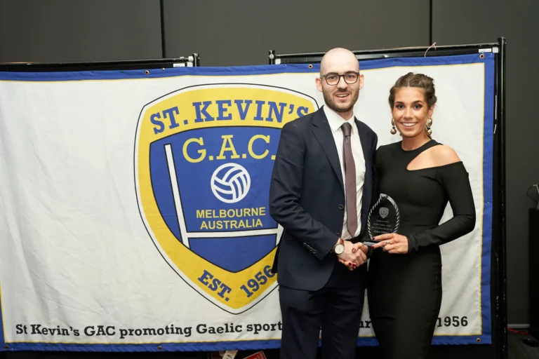 This image captures a celebratory moment at St. Kevin's 65th Anniversary Ball at Marvel Stadium, where a woman in a black one-shoulder dress holds a clear award trophy, smiling proudly. A man in a dark suit and tie stands beside her, congratulating her on the achievement. They are positioned in front of a banner displaying the St. Kevin's G.A.C. Melbourne Australia emblem and “EST. 1956,” emphasizing the formal and prestigious nature of the milestone anniversary awards presentation.
