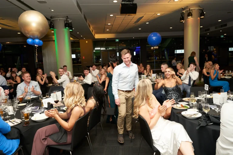 A male award recipient walks through guest tables at St. Kevin’s 65th Anniversary Ball at Marvel Stadium, smiling as attendees in formal attire clap and cheer. The ballroom is decorated with large gold and blue balloons, and tables feature dark tablecloths and elegant place settings. The image captures the celebratory energy and camaraderie of the evening as guests honor the achievements of their peers during the milestone anniversary celebration.