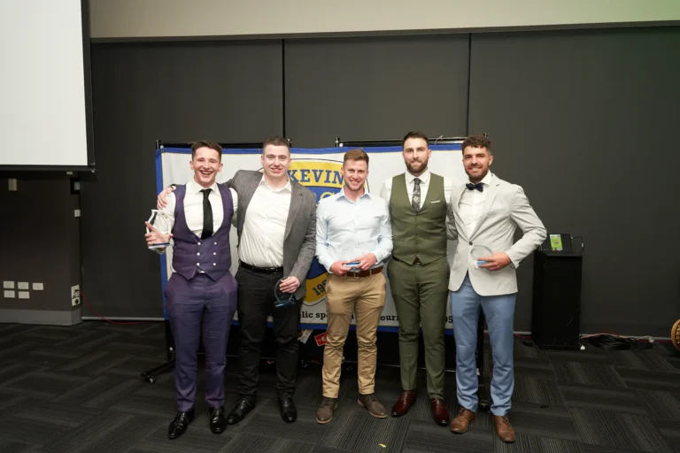 Group of award recipients at St. Kevin’s 65th Anniversary Ball, Marvel Stadium, Melbourne. Five men, dressed in formal and semi-formal attire, pose for a photograph, with two holding clear awards or trophies. A celebratory banner in the background references the milestone anniversary, while guest tables in the foreground hint at the elegant dinner setting and festive atmosphere of the event.