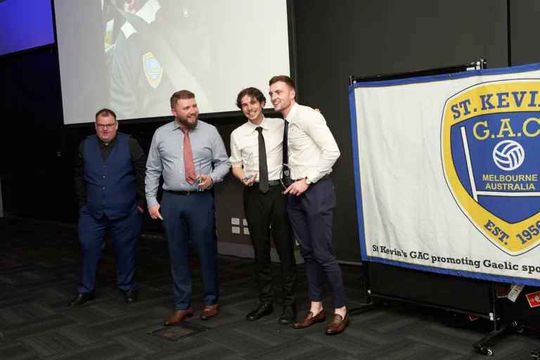 Four men celebrate at St. Kevin’s GAC 65th Anniversary Ball at Marvel Stadium, with three holding trophies and smiling warmly at the camera. Dressed in smart attire, they share a candid moment of achievement and camaraderie. A fourth man stands nearby, observing the group, while a banner in the background displays the St. Kevin’s GAC logo and highlights the club’s role in promoting Gaelic sports in Melbourne. A large screen behind them hints at the formal event setting. The image conveys joy, recognition, and festive spirit during this milestone anniversary celebration.