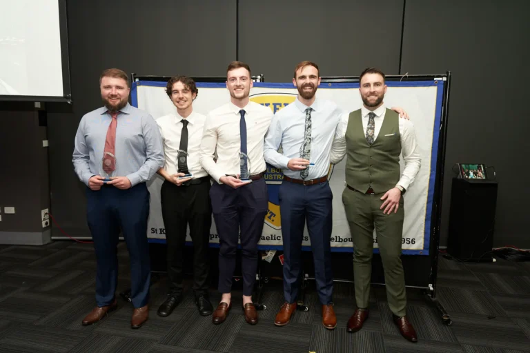 This image captures a celebratory moment at St. Kevin's 65th Anniversary Ball at Marvel Stadium, featuring five well-dressed men posing together. Four award winners, holding trophies or plaques, stand alongside a presenter or organizer, all smiling at the camera. Dressed in formal attire including shirts, ties, trousers, and a waistcoat, they exude camaraderie and pride. A large banner in the background highlights the official setting of the event, emphasizing the milestone celebration and the recognition of achievements during this significant anniversary ball.