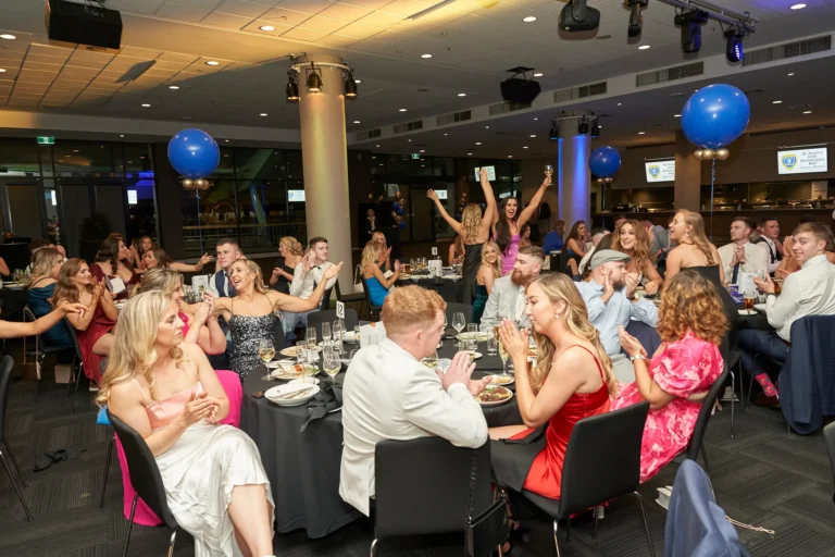 This wide-angle image from St. Kevin's 65th Anniversary Ball at Marvel Stadium captures a lively ballroom filled with elegantly dressed guests seated at round tables. Attendees engage in conversation, laughter, and applause during an awards presentation, with blue balloons adding a festive touch. The candid shot conveys genuine joy and camaraderie, highlighting the celebratory spirit as friends and colleagues cheer for award winners. The wide perspective emphasizes the scale and energy of the milestone anniversary event, reflecting the warmth and excitement shared among all present.
