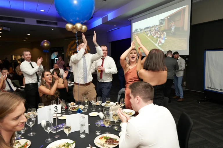 Wide-angle view of guests celebrating during the awards presentation at St. Kevin’s 65th Anniversary Ball at Marvel Stadium, Melbourne. Elegantly dressed attendees are seated at round tables, applauding and cheering as friends receive awards. The tables are set with wine glasses and plates, while large blue and gold balloons add a festive touch. In the background, a screen and a banner with the St. Kevin’s G.A.C. logo highlight the club’s achievements, capturing the lively camaraderie and joyous atmosphere of the milestone event.