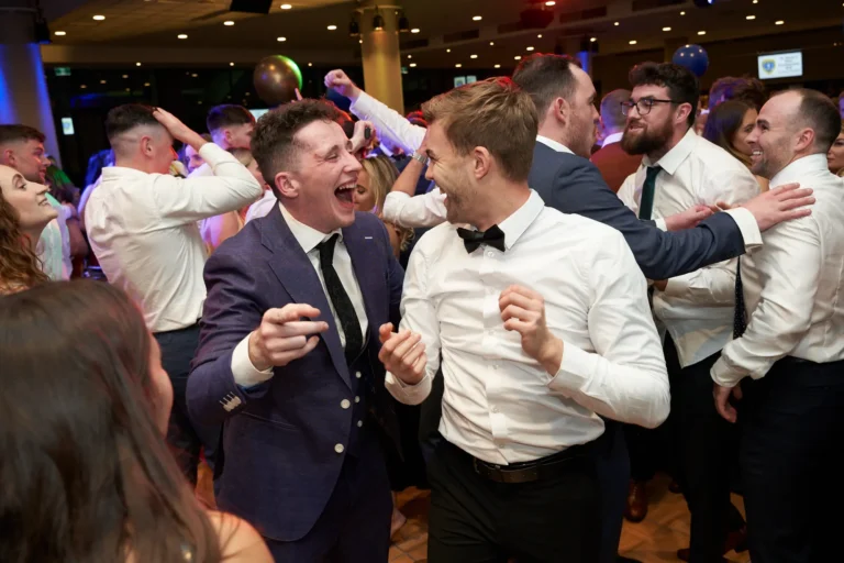 Candid moment at St. Kevin’s 65th Anniversary Ball at Marvel Stadium, capturing guests enjoying the dance floor. In the foreground, two men in formal attire—a blue suit and tie, and a white shirt with black bow tie—laugh and gesture energetically, sharing a joyful moment. Other guests surround them, smiling and interacting, while the brightly lit hall enhances the festive gala atmosphere. The image conveys lively celebration, camaraderie, and the spirited enjoyment of attendees at this milestone anniversary event.